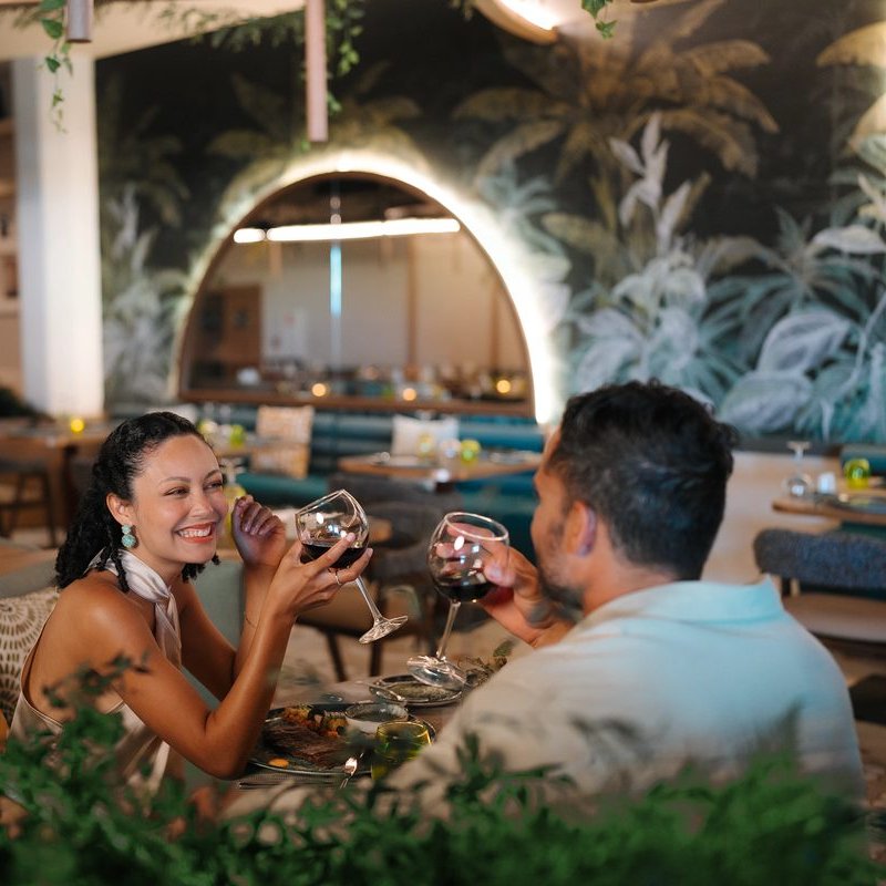 A couple laughs together over dinner, raising glasses across the table.