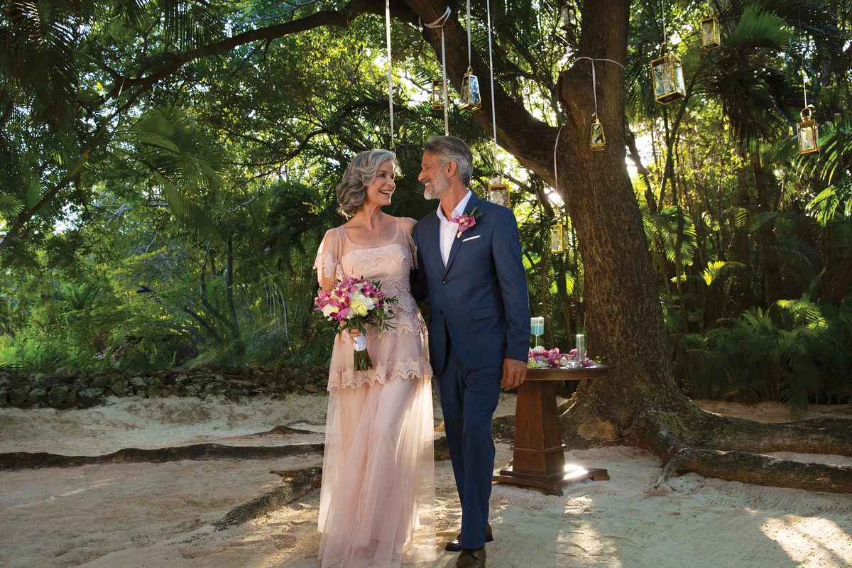 A silver-haired couple in formal attire stand close together on a beach beneath a tree hung with lanterns, she holding a bouquet
