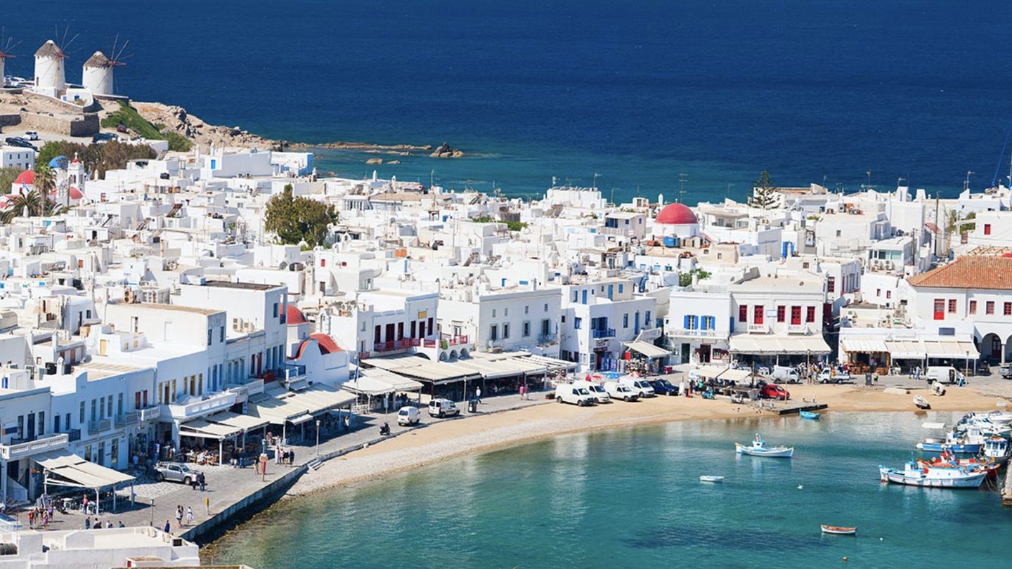 Whitewashed waterfront buildings and windmills curve around a small harbor of turquoise water