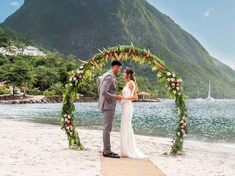 A couple holds hands beneath a floral arch on a white-sand beach, with a steep green mountain rising behind them