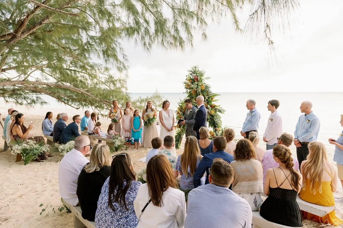 Guests are seated outdoors along a beachfront aisle as a couple celebrates their wedding ceremony
