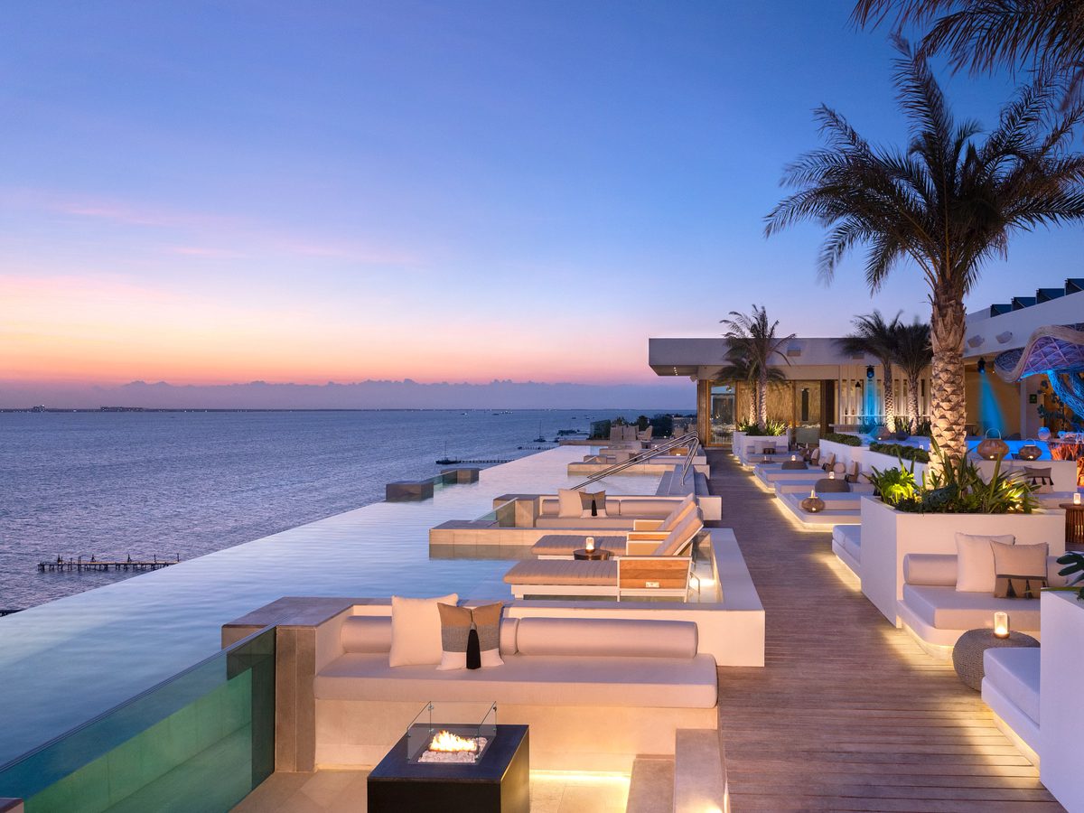 A seaside terrace at dusk with an infinity-edge pool, white lounge seating and a fire feature
