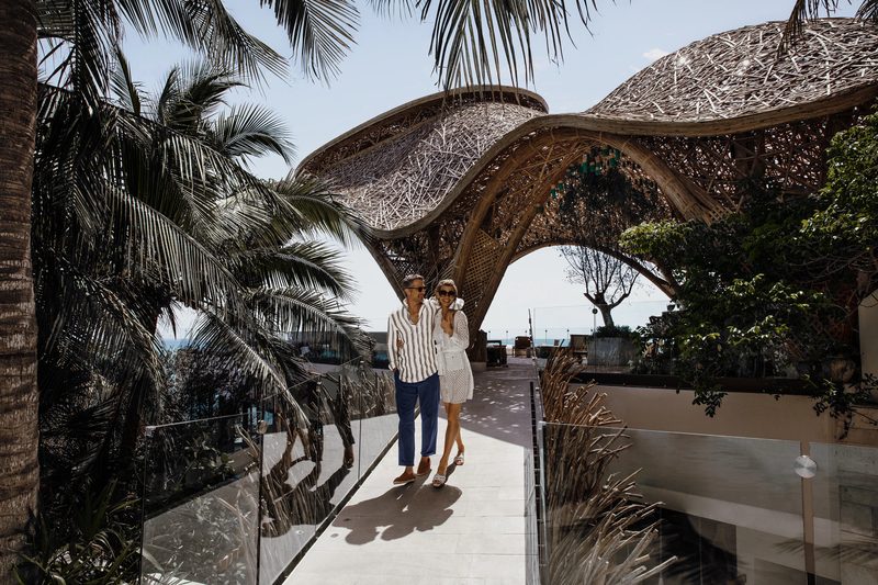 A couple walks along a sunlit path beneath a sculptural woven-timber canopy, surrounded by palms