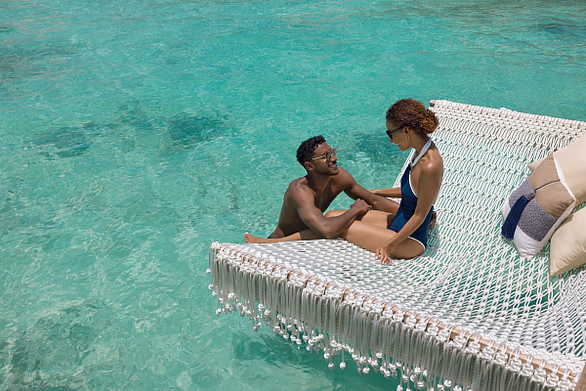 A couple relaxes together on a net hammock suspended over clear turquoise water