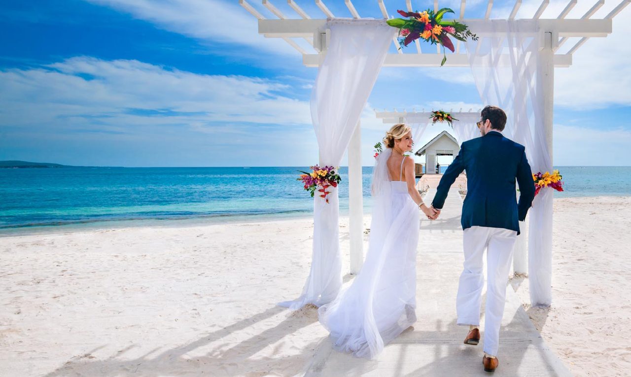 A bride and groom, seen from behind, walk hand in hand beneath a white draped pergola on a sunlit beach